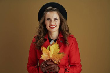 Merhaba Ekim. Düşünceli, orta yaşlı, kırmızı ceketli, siyah bereli, deri eldivenli, sarı sonbahar akçaağaç yapraklı, bej arkaplanlı bir kadın..