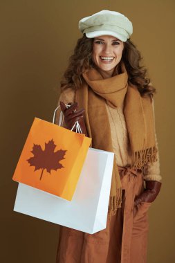 Merhaba Kasım ayı. Kahverengi arka planda izole edilmiş deri eldivenli ve kağıt çantalı 40 yaşında mutlu stil sahibi bir kadın..