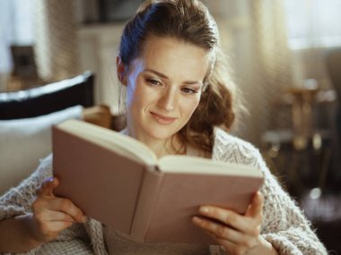 Güneşli bir sonbahar gününde, modern bir evde örülmüş, rahat bir hırka içinde, kitap okuyan gülümseyen genç bir kadın..
