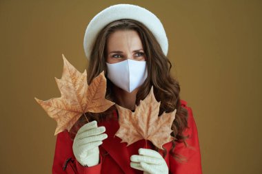 Merhaba sonbahar. Kırmızı paltolu, sarı sonbahar akçaağaç yaprağıyla düşünceli, şık ev hanımının portresi bej üzerinde izole edilmiş mesafeye bakıyor..