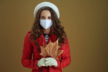 Koronavirüs salgını sırasında hayat. Kahverengi arka planda sarı sonbahar akçaağaç yaprağı olan kırmızı ceketli şık bir kadının portresi..
