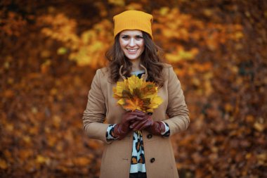 Merhaba Eylül. Sonbahar sarı yapraklı bej ceketli ve turuncu şapkalı gülümseyen genç kadın sonbaharda şehir parkında yaprak döküyor..