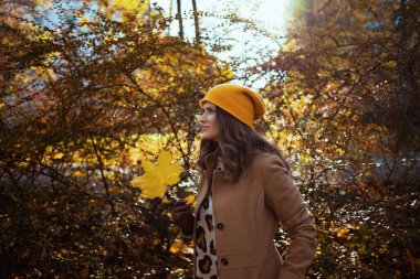 Merhaba Eylül. Sonbaharda şehir parkının dışındaki güz sarısı yapraklı kahverengi ceketli ve sarı şapkalı düşünceli modern kadın..