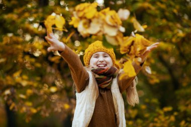 Merhaba Eylül. Kahverengi kazaklı ve turuncu şapkalı gülümseyen genç kız sonbaharda şehir parkında sonbahar yapraklarını fırlatıyor..