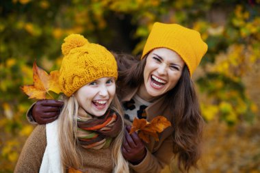 Merhaba Ekim. Sonbahar sarısı yapraklı turuncu şapkalı mutlu anne ve çocuk sonbaharda şehir parkında eğleniyor..