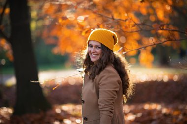Merhaba Eylül. Sonbaharda şehir parkında kahverengi ceketli ve sarı şapkalı gülümseyen genç bir kadın..