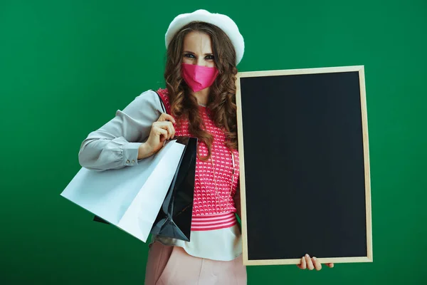 Covid 19パンデミックの間の生活 ピンクのノースリーブシャツにピンクの医療用マスクとショッピングバッグのスタイリッシュな女性は 緑の背景に空白の黒いボードを示す — ストック写真