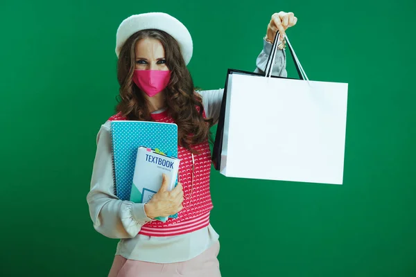 コロナウイルスのパンデミックの間の生活 教科書 ノート ピンクの医療マスクや緑の背景にショッピングバッグとピンクの袖のないシャツの若い女性の肖像画 — ストック写真