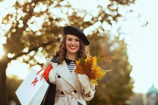 Hello autumn. smiling modern middle aged woman in beige trench coat with shopping bags and autumn yellow leaves outdoors on the city street in autumn.