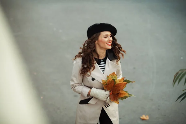 ベージュのトレンチコートと秋の黄色の葉と黒のベレー帽で幸せな流行の女性の上のビュー秋に街の通りに屋外 — ストック写真