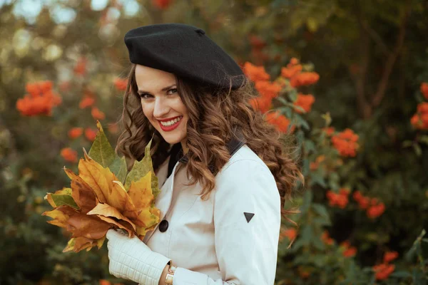 今年秋天 穿着米黄色风衣 头戴黑色贝雷帽 头戴秋天黄色叶子的40岁的年轻女性在城外的城市街道上微笑着 — 图库照片