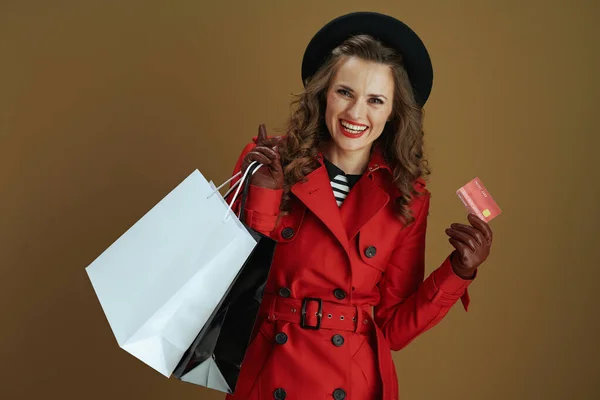 9月だ 赤コートとクレジットカードで黒ベレー帽でスタイリッシュな40歳の女性の笑顔の肖像画 革の手袋と茶色の背景に隔離された紙のショッピングバッグ — ストック写真