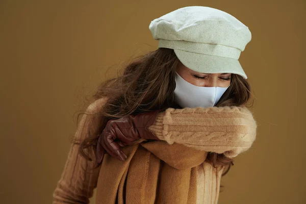 Hello autumn. trendy woman in scarf with white medical mask sneezing into elbow against brown background.