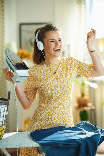 現代の笑顔40歳の主婦黄色のドレスアイロン台で晴れた日に現代のリビングルームでヘッドフォンで音楽を聴きながら — ストック写真