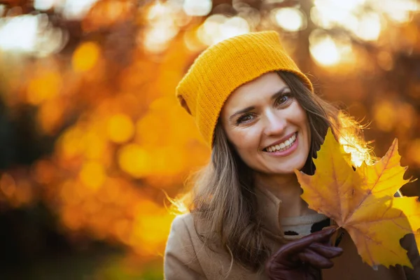 Hello autumn. happy stylish middle aged woman in beige coat and orange hat with autumn yellow leaves outside on the city park in autumn.