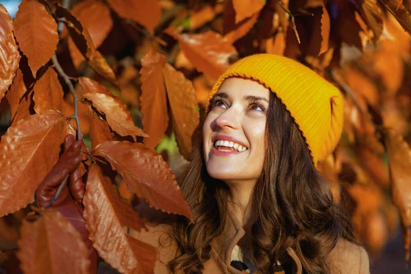 秋の都会の公園の外の紅葉の中で現代女性の茶色のコートと黄色の帽子を笑って — ストック写真
