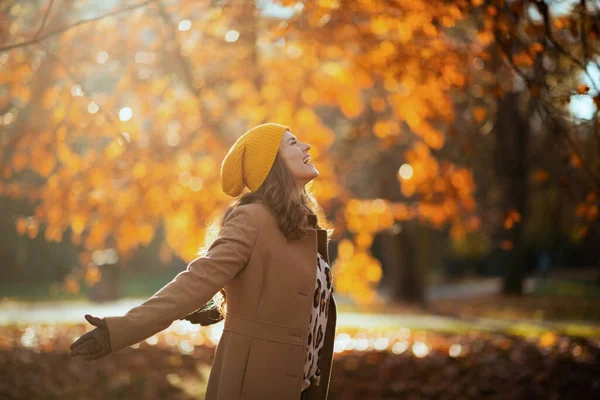 Merhaba sonbahar. Sonbaharda şehir parkında bej ceketli ve turuncu şapkalı mutlu orta yaşlı kadın neşe saçıyor..