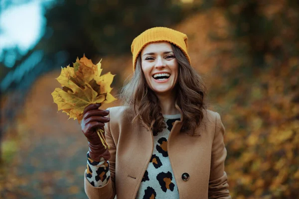 10月だ 秋に市公園の屋外で秋の黄色の葉を持つ茶色のコートと黄色の帽子の幸せなエレガントな女性 — ストック写真