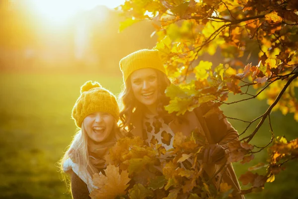 オレンジ色の帽子をかぶった現代の母親と子供の笑顔秋の公園の屋外の紅葉の間で — ストック写真