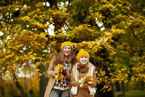 秋には公園の外に黄色の葉を持つ黄色の帽子の若い母親と娘を笑顔で — ストック写真