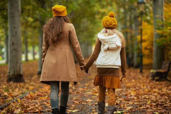 秋には公園の外のオレンジの帽子の現代の母親と娘の後ろから見た遊歩道を楽しんで — ストック写真
