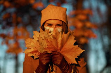 Merhaba Ekim. Kahverengi ceketli, sarı şapkalı, tıbbi maskeli ve sonbahar sarısı yapraklı mutlu bir kadın sonbaharda şehir parkında..