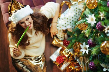 Merry Christmas. Upper view of smiling elegant woman in gold sequin skirt and sweater near Christmas tree and gift boxes eating celery.