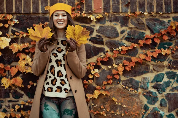 Hello autumn. happy modern middle aged woman in brown coat and yellow hat with autumn yellow leaves against stone wall outdoors on the city street in autumn.