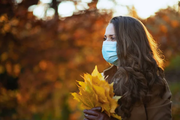 9月だ 秋に市公園の外で医療マスクと秋の黄色の葉を持つベージュのコートで高価なスタイリッシュな中年女性 — ストック写真