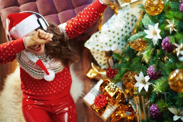 圣诞快乐 身穿红毛衣 头戴圣诞礼帽的快乐年轻女子 头戴耳机听音乐 在圣诞树和礼品盒前做涉猎手势的上图 — 图库照片