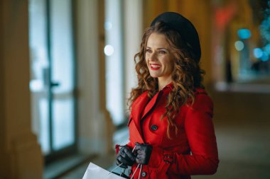 Kış eğlencesi. Kırmızı paltolu ve siyah bereli mutlu genç bir turist. Alışveriş çantalı. Geceleri dışarıdaki dükkanın camına bakıyor..