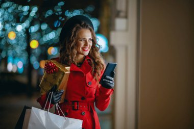Winter Fun. smiling young woman shopper in red coat and black beret with shopping bags, gifts and smartphone looking at the shop window and sending text message outdoors in the city street at night.