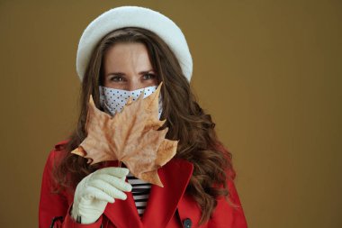 Koronavirüs salgını sırasında hayat. Kahverengi arka planda sarı sonbahar akçaağaç yaprağı olan kırmızı ceketli modaya uygun kadın portresi..