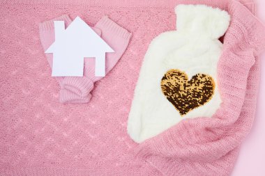 winter flat lay with mittens, hot water warmer and paper house on pink background.