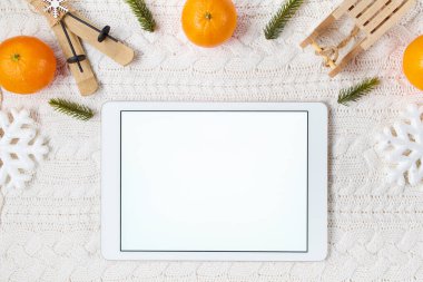winter flat lay with tablet pc, snowflakes, mandarins, sleds and skis on white knitted background.