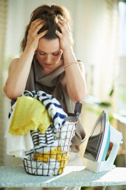 Stresli 40 yaşındaki ipek bluz ve buharlı bej pantolonlu ev hanımı, güneşli bir günde modern evde çamaşır sepeti ve ütü masası..