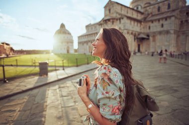 Cattedrale di Pisa yakınlarında, çiçekli, klasik fotoğraf makinesi ve sırt çantalı mutlu orta yaşlı bir gezgin kadın..