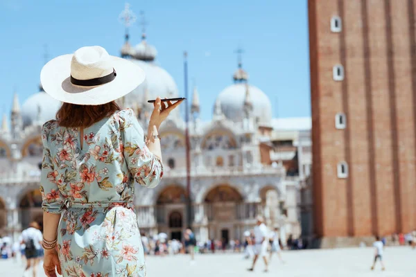 花のドレスを着た流行の観光客の女性の後ろから見た帽子はヴェネツィア イタリアのサンマルコ広場でスマートフォンで話しています — ストック写真