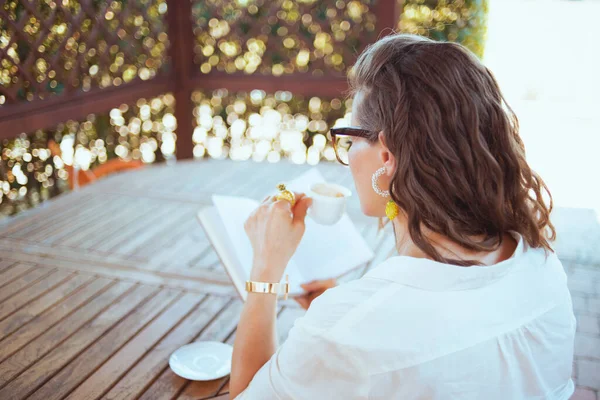从后面看穿着白衬衫 眼镜坐在桌旁喝咖啡 在阳台看书的女人 — 图库照片