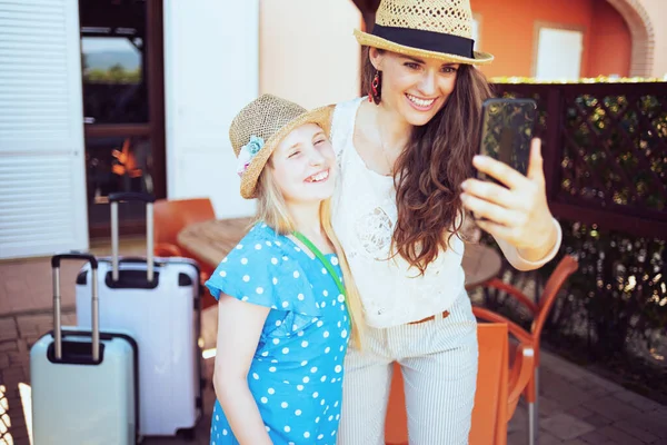 ホテルのテラスでビデオ通話をしている車のバッグを持つ帽子の流行の母親と娘を笑顔で — ストック写真