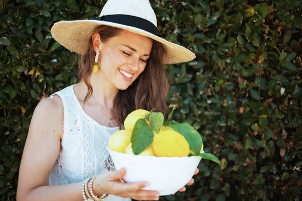 白いシャツに地元の農場のレモンのプレートと緑の壁の近くの屋外の帽子を持つエレガントな女性の笑顔 — ストック写真