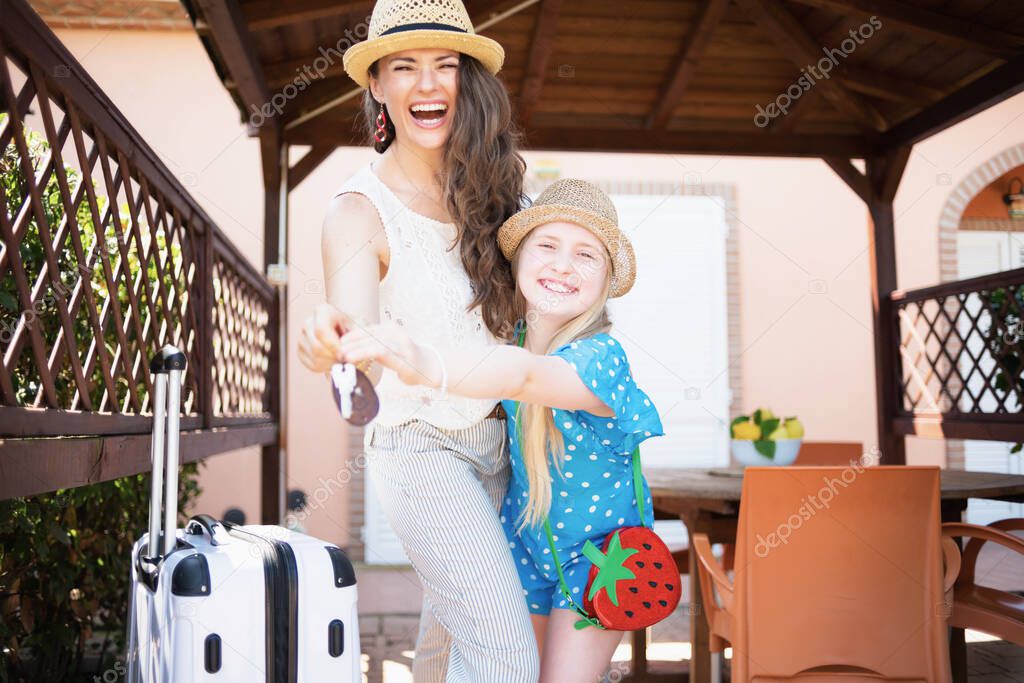 happy stylish mother and child in hats with wheel bags and keys in the patio of guest house hotel.