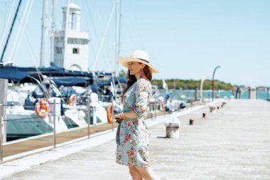 Gülümseyen modaya uyan, çiçekli elbiseli, şapkalı, iskelede gezen kadın..