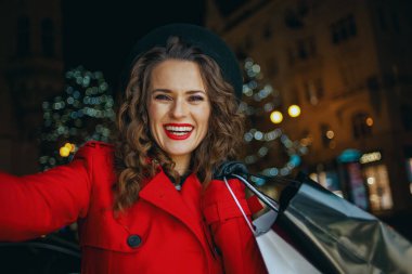 Kış eğlencesi. Kırmızı paltolu ve siyah bereli gülümseyen modern yalnız turist kadın portresi. Alışveriş torbaları ile geceleri şehrin dışında selfie çekiyor..