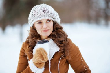 Gülümseyen 40 yaşında, eldivenli ve elinde çay fincanı olan, örgü şapkalı ve koyun derisi ceketli bir kadın kışın şehir parkında..