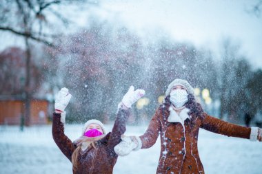 Örgü şapkalar ve eldivenli kuzu derisi paltolar ve örgü şapkalı ve kuzu derisi ceketli modern anne ve kız kışın şehir parkında neşe içinde eğleniyorlar..