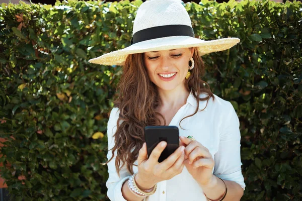 緑の壁の近くの屋外でスマートフォンアプリを使用して帽子を持つ白いシャツの幸せな中年主婦 — ストック写真