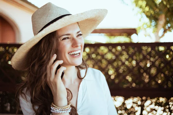 在宾馆的阳台上 身穿白衬衫 头戴礼帽 头戴智能手机 面带微笑的时髦女性 — 图库照片