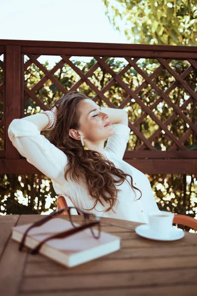 relaxed elegant woman in white shirt with cup of coffee, book and eyeglasses sitting at the table in the patio of guest house hotel.