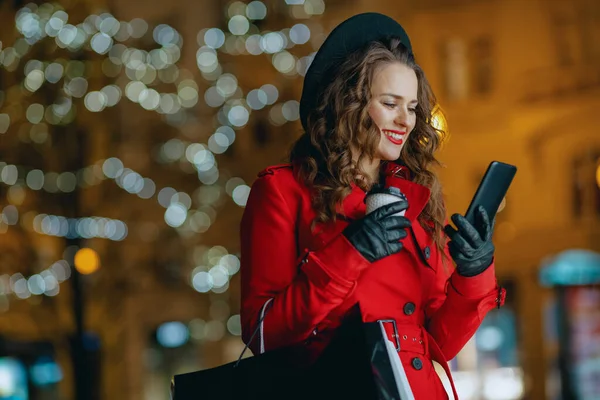 冬の楽しみ 幸せな中年女性買い物客で赤コートと黒ベレー帽でショッピングバッグ カップのホットチョコレートとスマートフォンオンラインで市内の外で夕方 — ストック写真